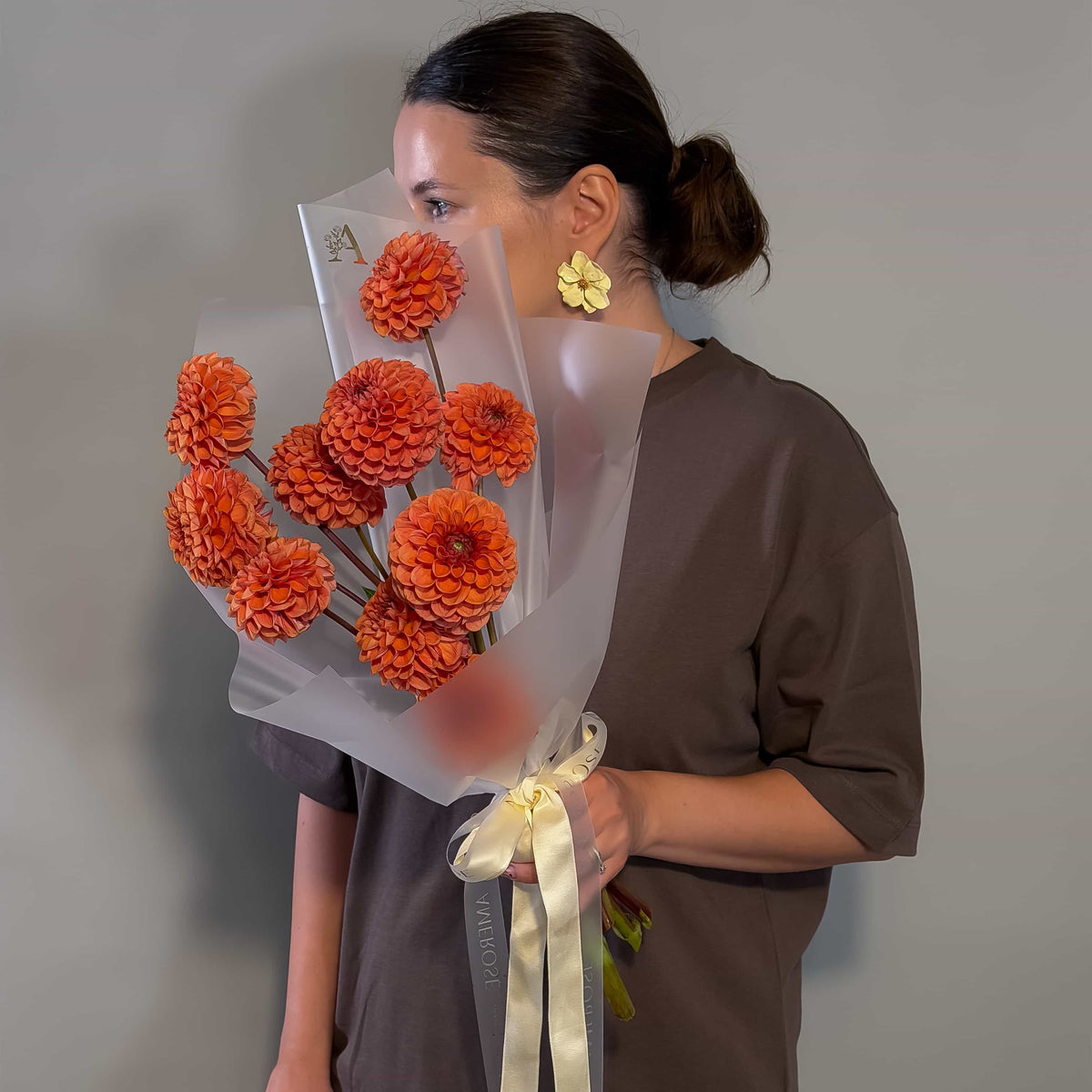 A woman in a brown t-shirt holds a bouquet of bronze dahlias wrapped in translucent white paper with a cream ribbon, standing against a plain gray background.
