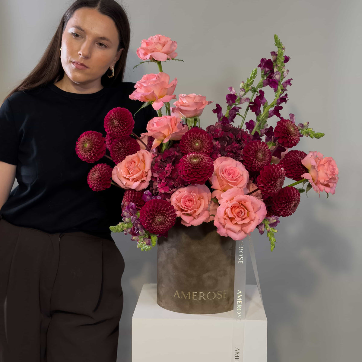 A woman in a black top and dark trousers stands beside a large floral arrangement in a taupe box labeled AMEROSE, set on a white pedestal against a light gray wall.