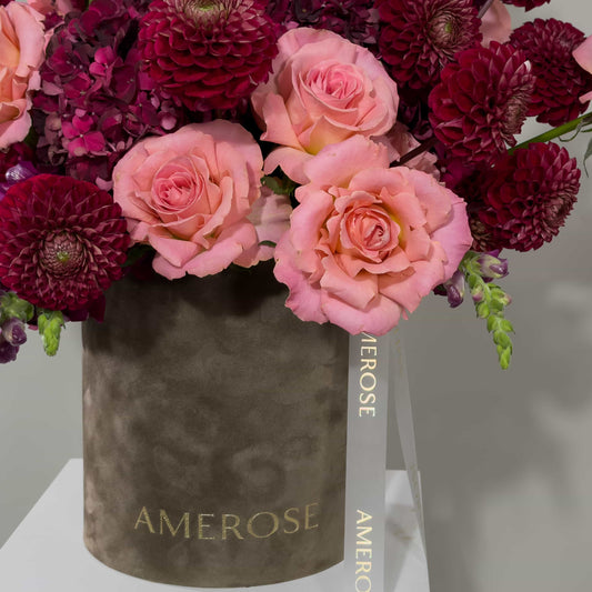 A floral arrangement of pink roses and deep burgundy dahlias in a taupe cylindrical box with gold 'AMEROSE' lettering and a white hanging ribbon.