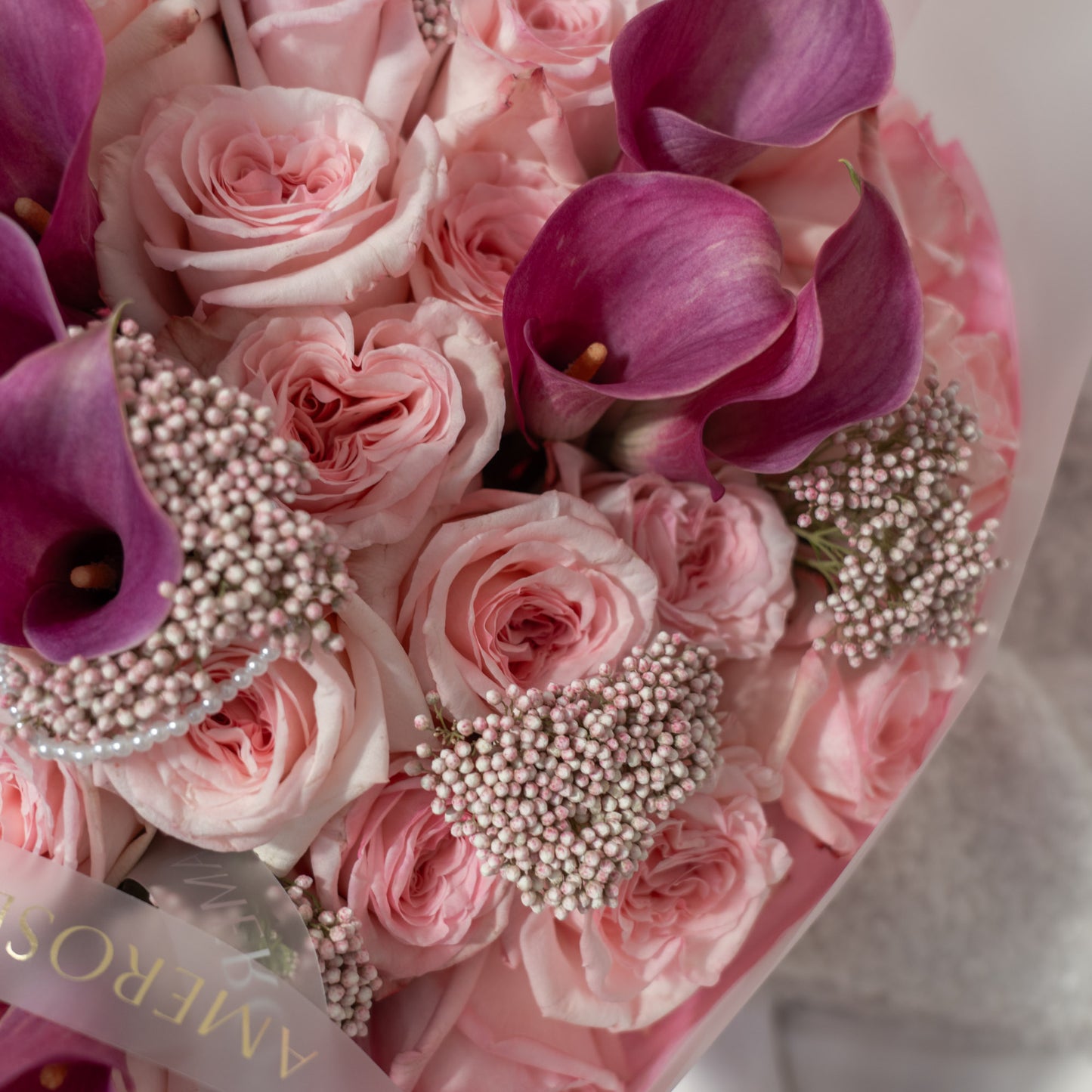 Top-down close-up of blush pink roses, purple magenta mini calla lilies and pearl-like ivory rice flowers with AMEROSE ribbon — luxury bouquet detail London