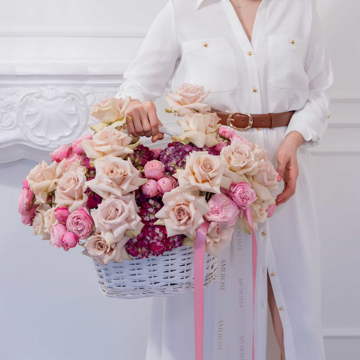 A women holding a romantic basket with hydrangea, nude roses and pink spray roses against a white wall.