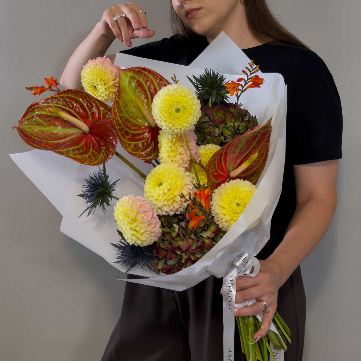 A person holding a large, vibrant floral bouquet wrapped in white paper. The bouquet features red and green anthuriums, yellow dahlia-like flowers, foliage, and hydrangeas tied with a white ribbon printed with AMEROSE.