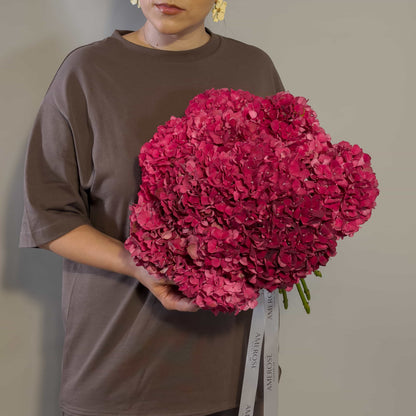 A person wearing a brown oversized T-shirt holds a large bouquet of burgundy hydrangea flowers with white ribbons against a plain light gray background.