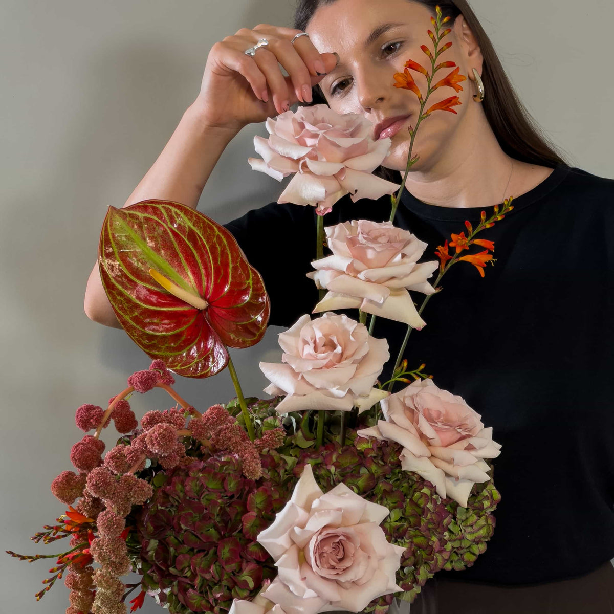 A woman in a black top holds a large, vibrant floral arrangement featuring pale pink roses, a red and green anthurium, and textured burgundy flowers against a neutral gray background.