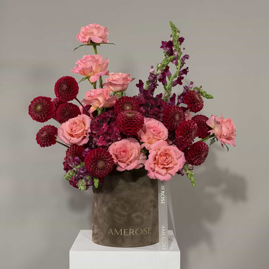 A floral arrangement of pink and deep burgundy flowers in a taupe cylindrical vase with the brand name AMEROSE printed on it, displayed on a white pedestal against a neutral gray background.