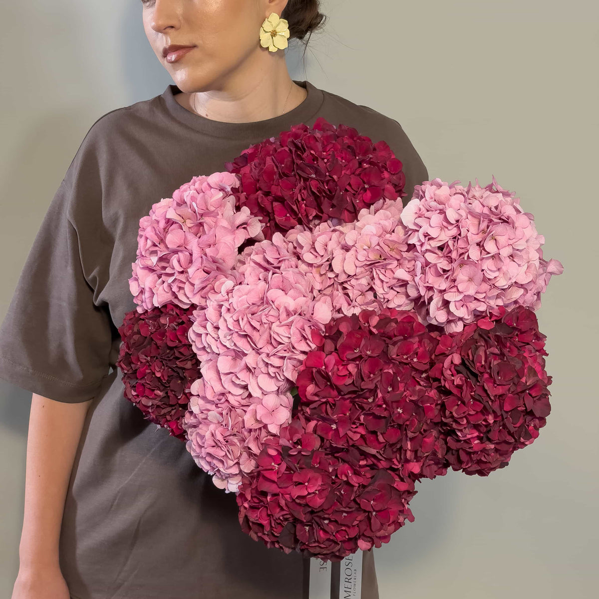 A person wearing a brown t-shirt holds a large bouquet of fresh cut hydrangeas in shades of pink and deep burgundy against a plain gray wall.