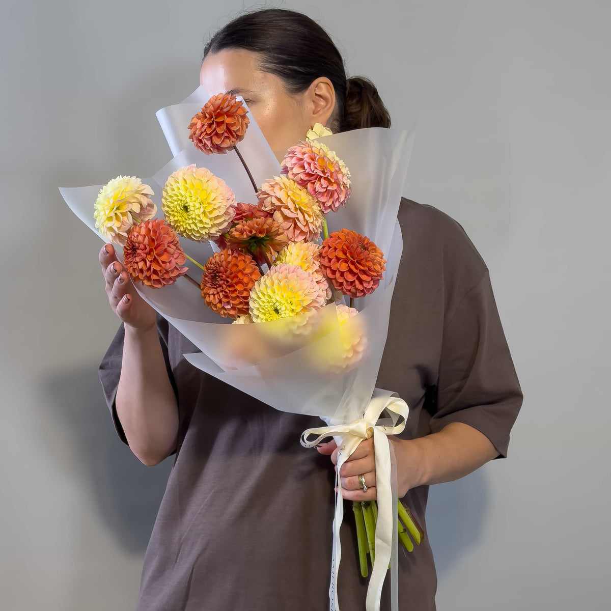 A person holding a bouquet of large dahlia flowers in orange, yellow, and peach tones, wrapped in translucent white paper with a cream ribbon, against a plain gray background.