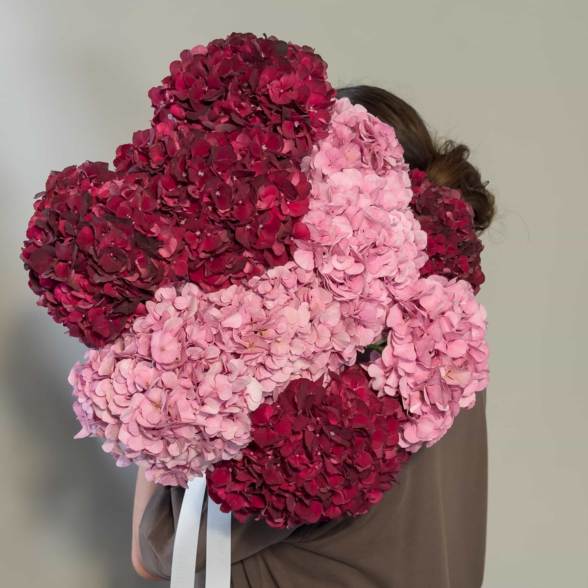 A person from behind holding a large bouquet of fresh cut hydrangea flowers in dark red and light pink shades, wearing a brown top against a plain light gray background.
