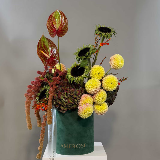 A floral arrangement in a dark green velvet vase with the brand name 'AMEROSE' printed in gold. The arrangement features yellow dahlia flowers, dark sunflowers, reddish-brown anthuriums, and hydrangeas, displayed on a white pedestal against a neutral gray background.