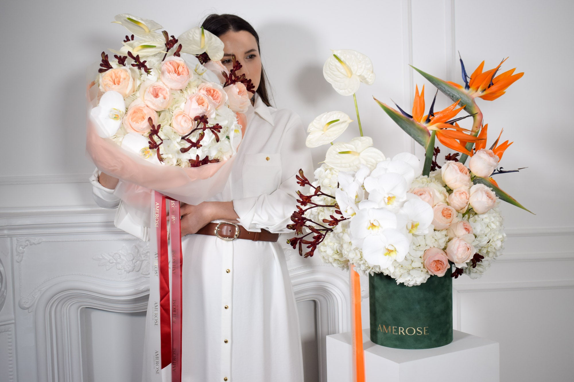 Woman holding a bouquet of flowers next to a hatbox arrangement by AMEROSE against a white wall.