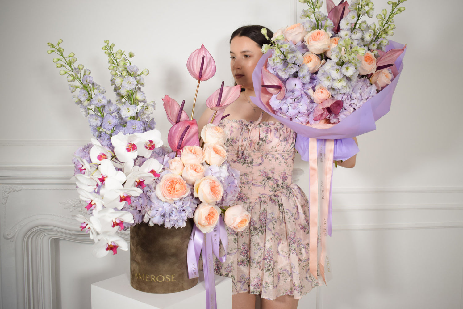 Woman holding a bouquet of flowers next to a large floral arrangement by AMEROSE against a white background.