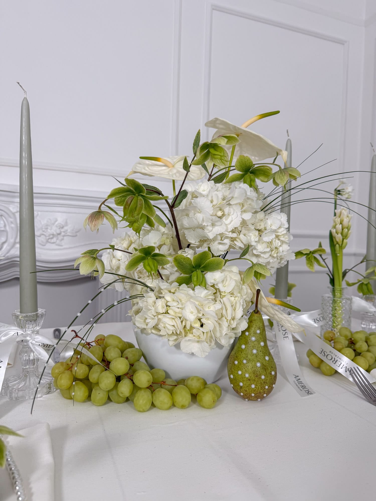 Decorative romantic table setting with white and green flowers, green grapes, and a pear on a white surface by AMEROSE London for corporate or private events.