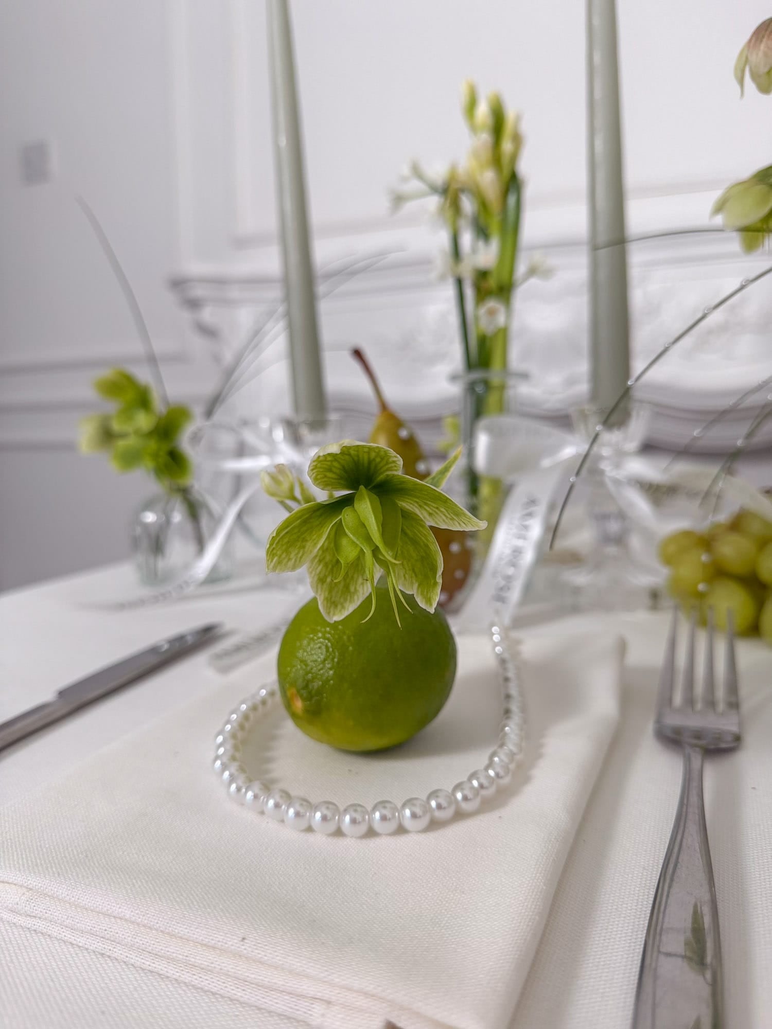 Decorative table setting with a green fruit and pearl necklace on a white tablecloth by AMEROSE London.