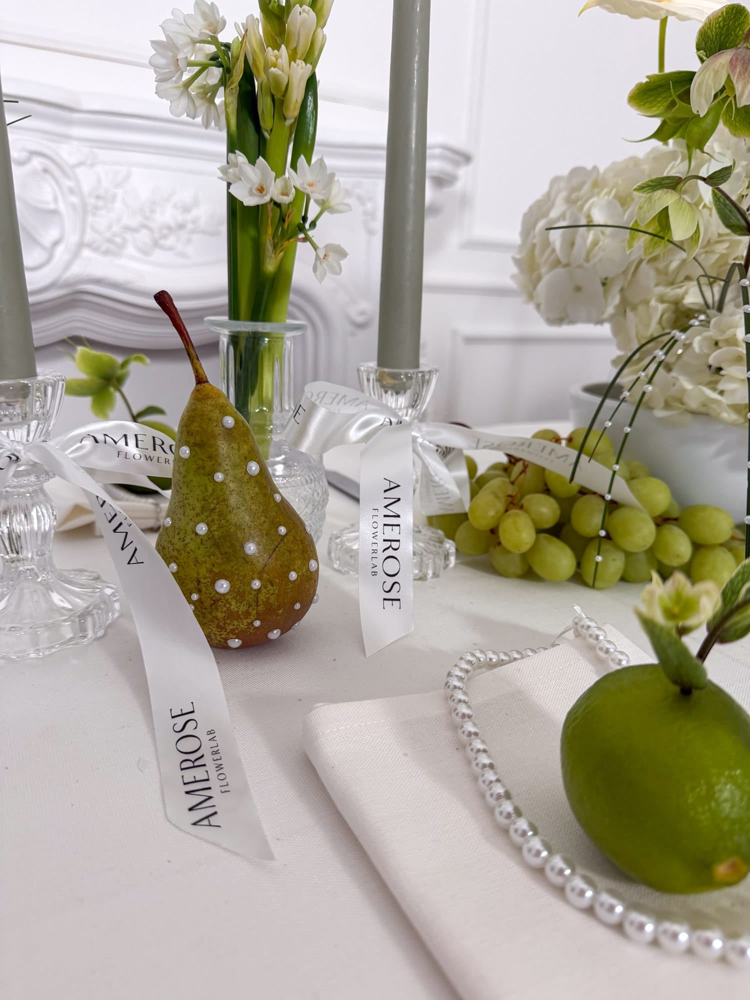 Decorative romantic table setting with pears, grapes, and flowers on a white tablecloth by AMEROSE London