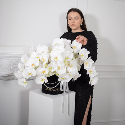 AMEROSE Blanc Orchids signature hatbox — dome of white phalaenopsis orchids in black velvet hatbox with pearl detail, held by model in black dress