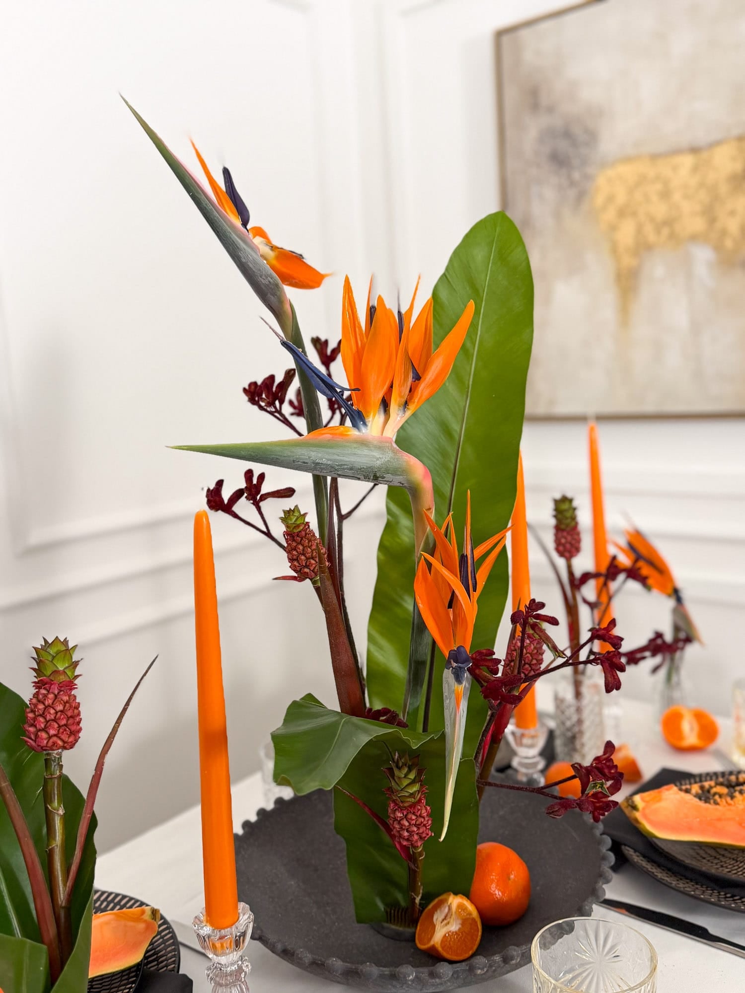 Decorative table setting with tropical flowers like strelitzia and ananas, candles, and fruit on a white surface by AMEROSE London.