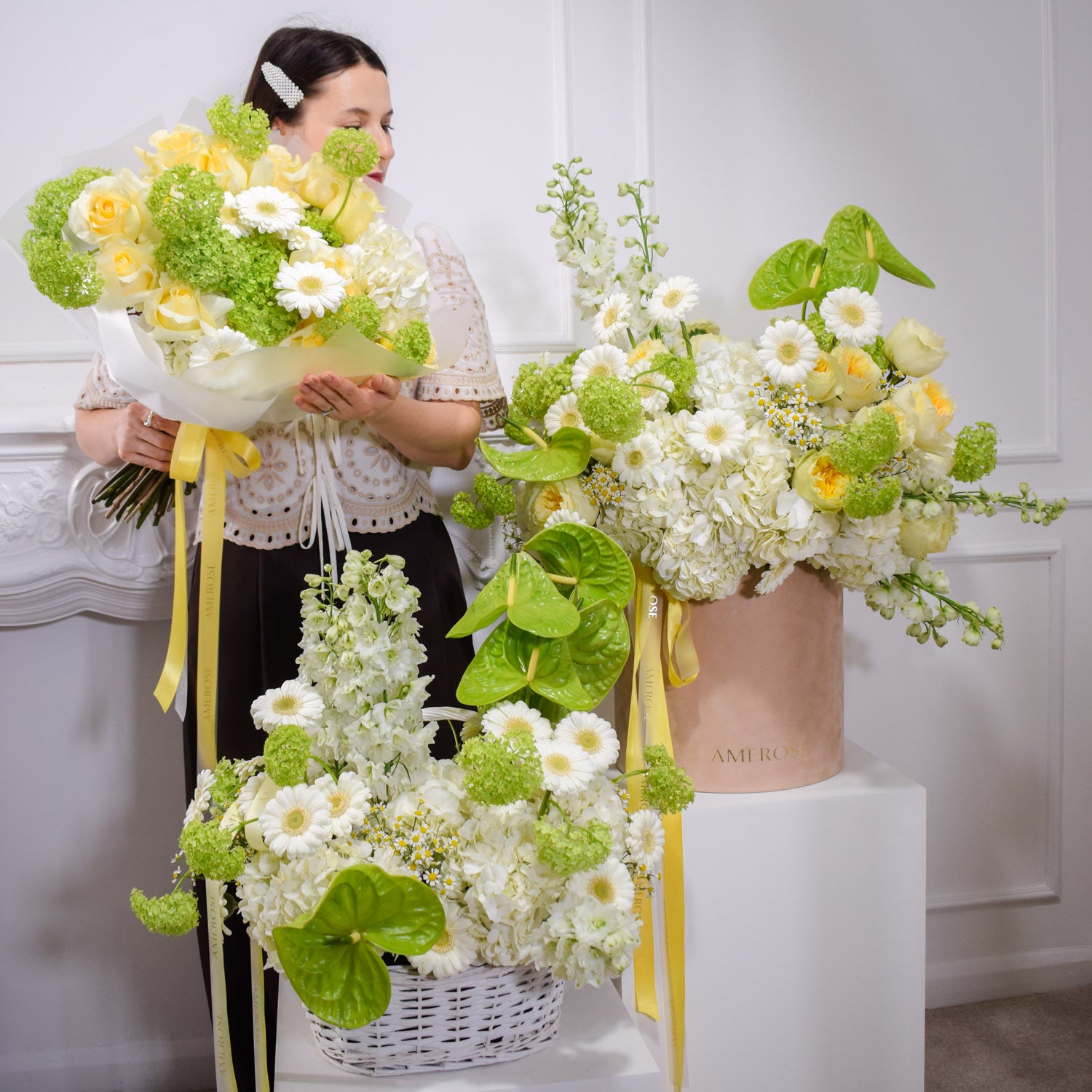Woman holding a bouquet of flowers with various floral arrangements by AMEROSE against a white wall.