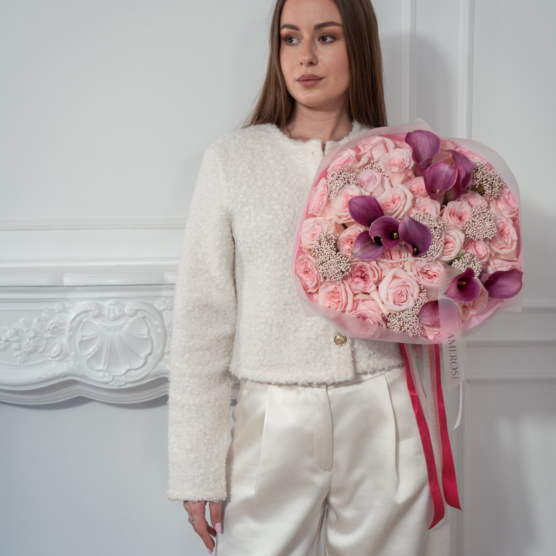Woman in white bouclé jacket holding a luxury bouquet of blush roses, purple calla lilies and rice flowers in pink tissue with bright pink AMEROSE ribbon — premium flower delivery London
