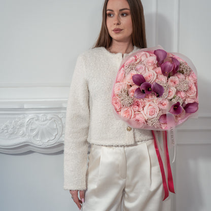Woman in white bouclé jacket holding a luxury bouquet of blush roses, purple calla lilies and rice flowers in pink tissue with bright pink AMEROSE ribbon — premium flower delivery London
