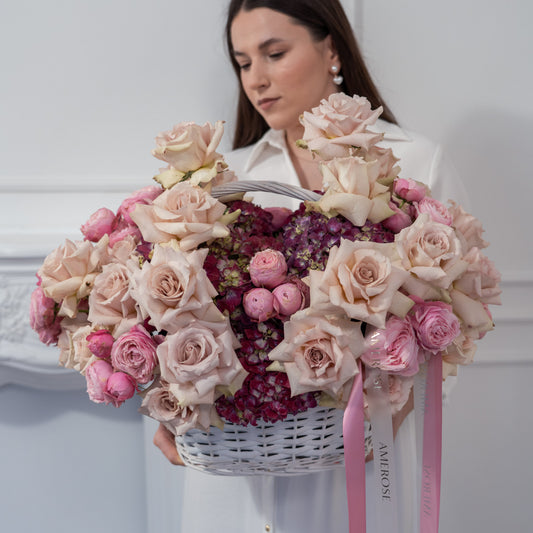 Woman holding an overflowing luxury flower basket with blush nude roses, pink spray roses and burgundy hydrangeas, tied with AMEROSE pink satin ribbon — made-to-order flower delivery London

