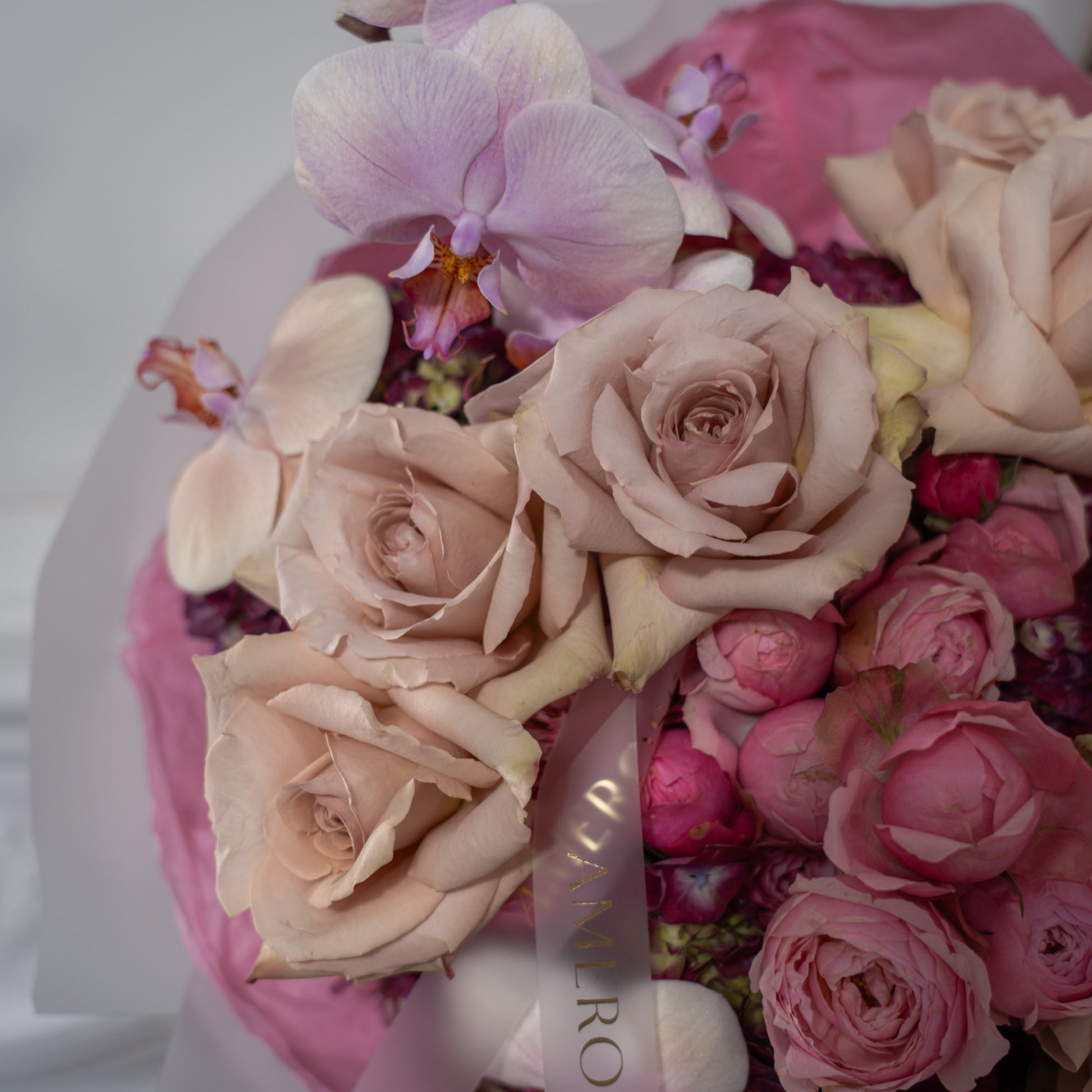 Close-up of dusty mocha roses, hot pink spray roses, lavender phalaenopsis orchid and burgundy hydrangeas with AMEROSE gold ribbon — luxury textured bouquet detail London