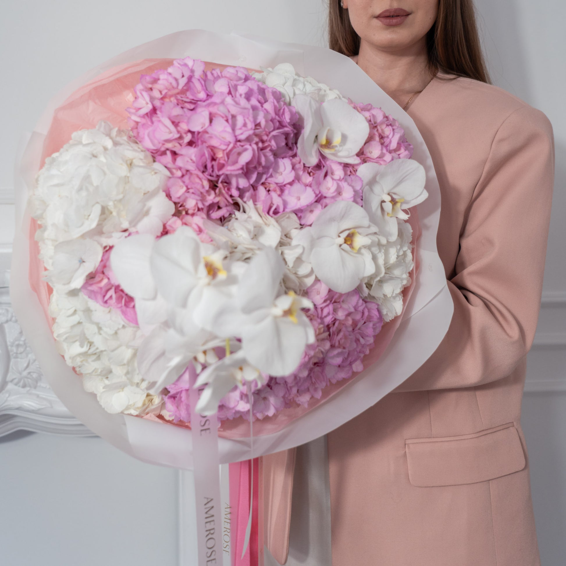 Woman in blush blazer holding an oversized luxury bouquet of pink hydrangeas and white phalaenopsis orchids elegantly wrapped with pink AMEROSE ribbon — premium flower delivery London