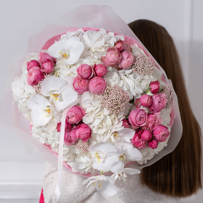 Woman in white bouclé jacket holding an oversized luxury bouquet of hot pink spray roses, white hydrangeas and orchids in pink tissue with fuchsia AMEROSE ribbon — premium flower delivery London
