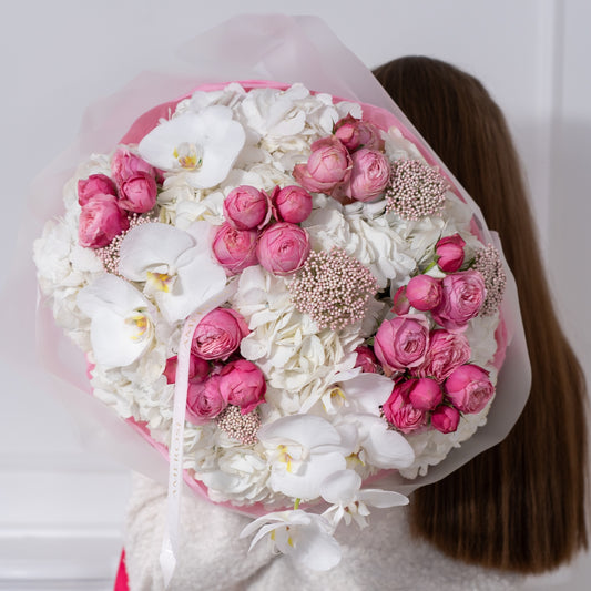 Woman in white bouclé jacket holding an oversized luxury bouquet of hot pink spray roses, white hydrangeas and orchids in pink tissue with fuchsia AMEROSE ribbon — premium flower delivery London
