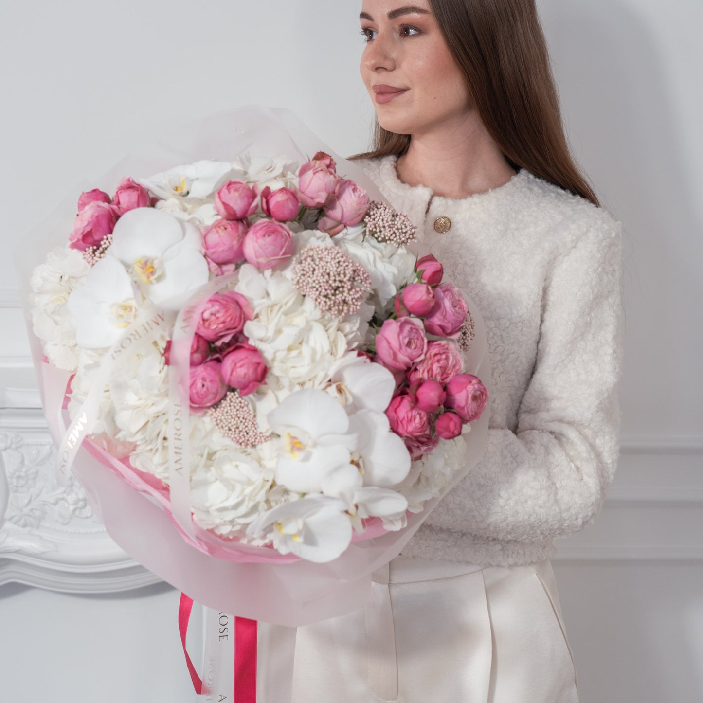 Woman in white bouclé jacket holding an oversized luxury bouquet of hot pink spray roses, white hydrangeas and orchids in pink tissue with fuchsia AMEROSE ribbon — premium flower delivery London

