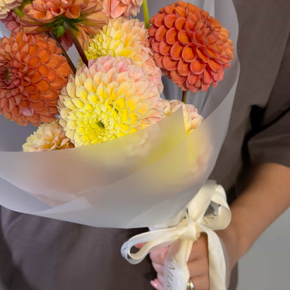 A close-up of a hand holding a bouquet of dahlia flowers wrapped in white paper. The flowers are in shades of orange, peach, and pale yellow, tied with a cream ribbon.