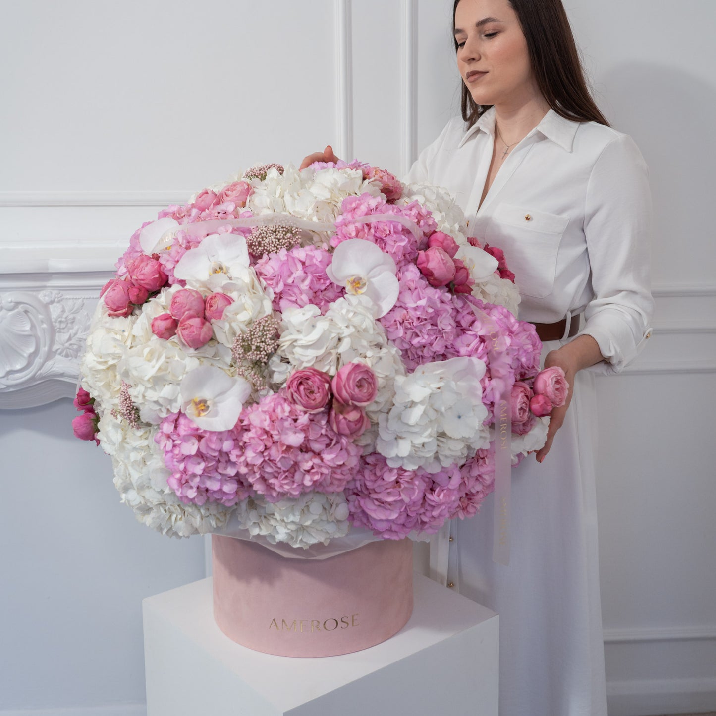 Woman standing beside an extraordinary large AMEROSE blush velvet hatbox overflowing with pink hydrangeas, white orchids and spray roses on white plinth — luxury flower delivery London