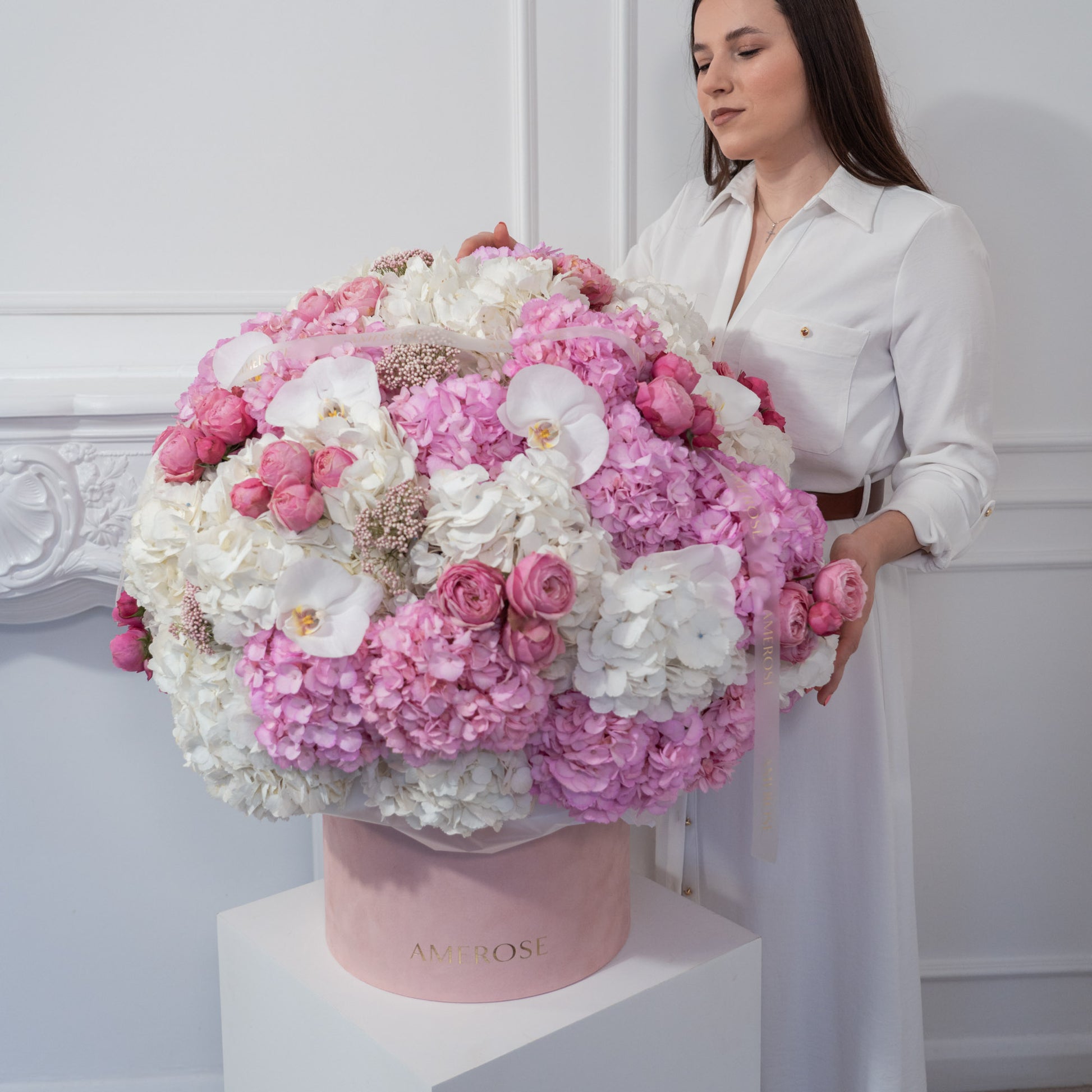 Woman standing beside an extraordinary large AMEROSE blush velvet hatbox overflowing with pink hydrangeas, white orchids and spray roses on white plinth — luxury flower delivery London