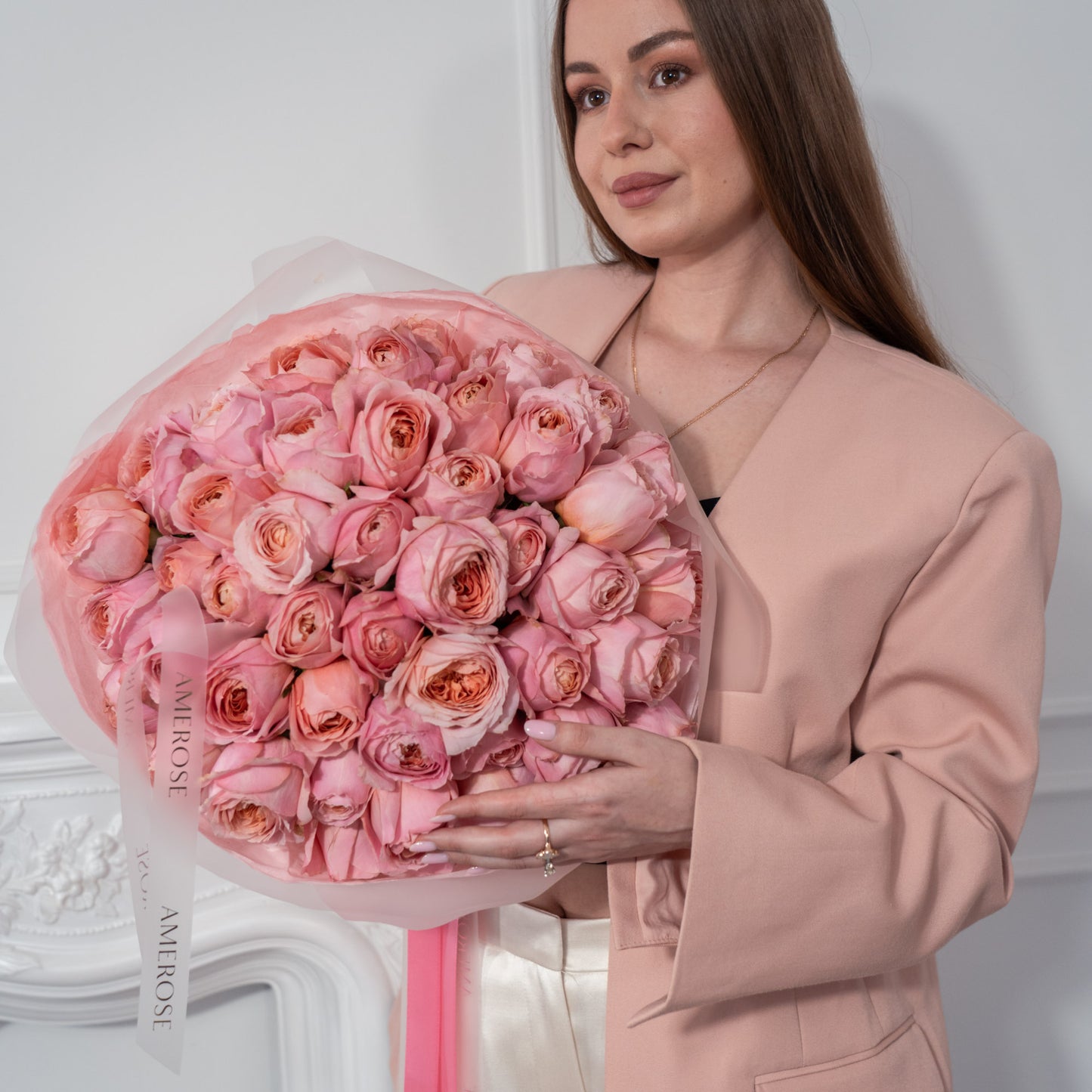 Woman in blush blazer holding a large luxury bouquet of pink garden roses wrapped in frosted film and peach tissue with AMEROSE pink ribbon — premium flower delivery London
