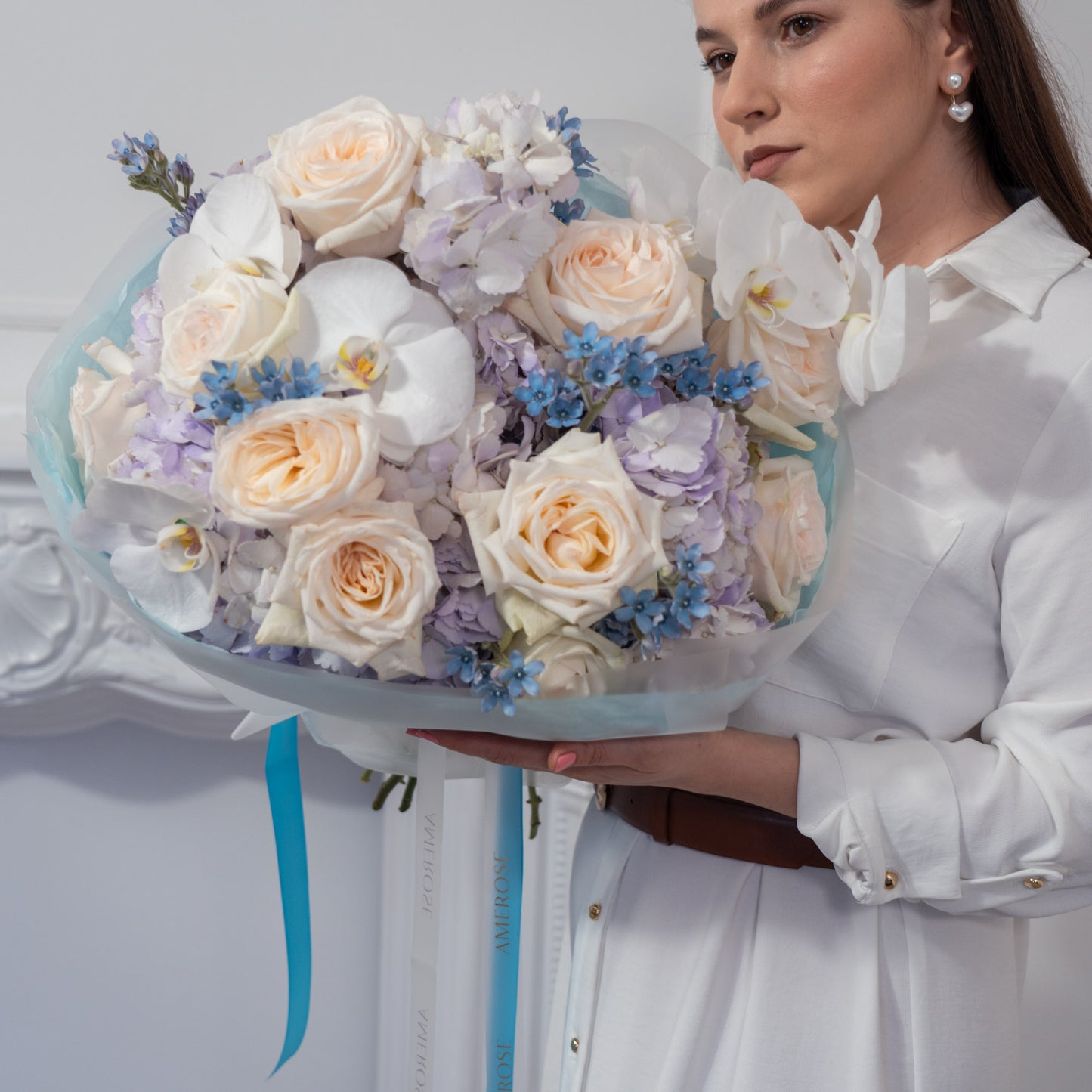 Woman holding a luxury bouquet of cream roses, white orchids, blue forget-me-nots and lavender hydrangeas wrapped in pale blue tissue with teal AMEROSE ribbon — premium flower delivery London