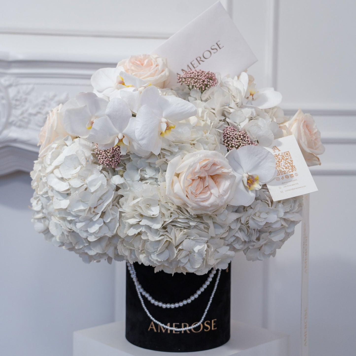 AMEROSE black velvet hatbox with double pearl chain handle, overflowing with white hydrangeas, blush garden roses, white orchids and pink wax flowers — luxury hatbox flowers London