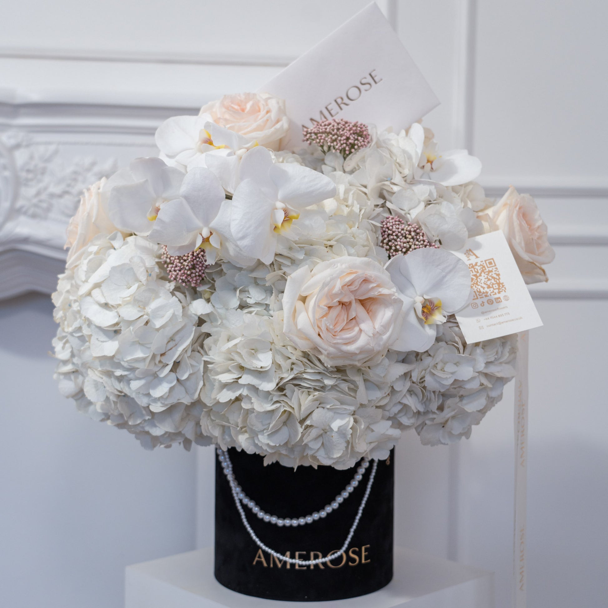 AMEROSE black velvet hatbox with double pearl chain handle, overflowing with white hydrangeas, blush garden roses, white orchids and pink wax flowers — luxury hatbox flowers London