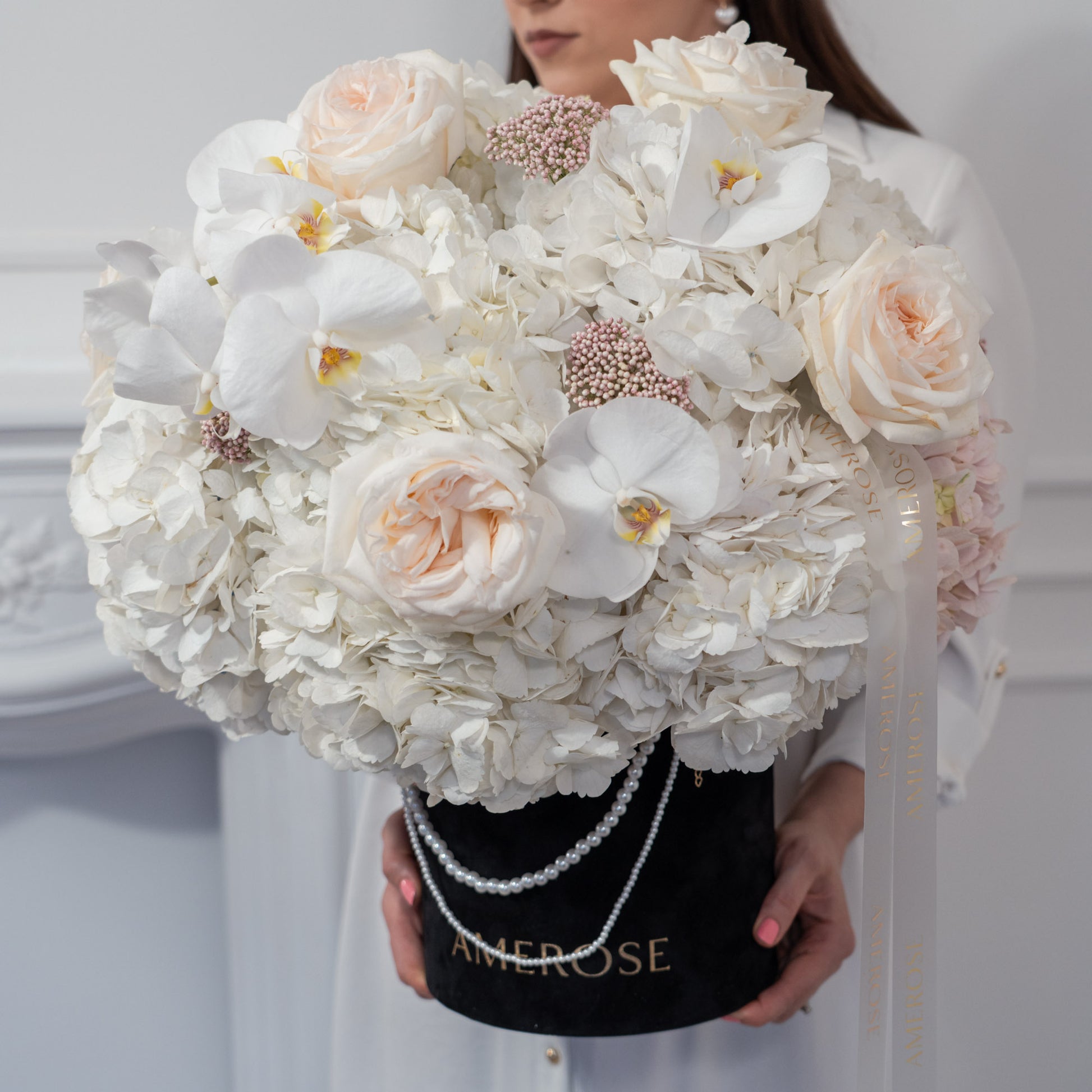 Woman holding an oversized AMEROSE black velvet hatbox with pearl handle, filled with white hydrangeas, blush roses and orchids — premium luxury flower delivery London