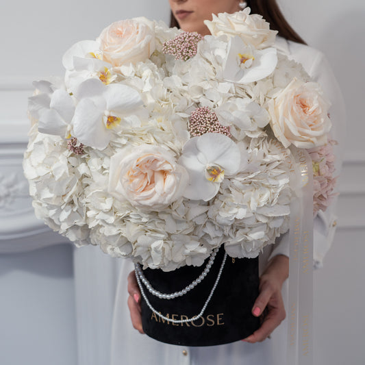 Woman holding an oversized AMEROSE black velvet hatbox with pearl handle, filled with white hydrangeas, blush roses and orchids — premium luxury flower delivery London