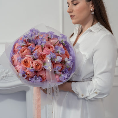 Woman holding a large luxury bouquet of coral peach roses and lavender sweet peas wrapped in lilac sheer tissue with AMEROSE ribbon — premium flower delivery London