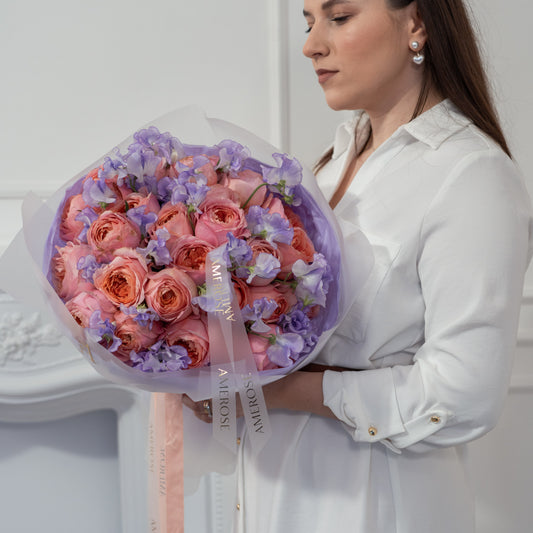 Woman holding a large luxury bouquet of coral peach roses and lavender sweet peas wrapped in lilac sheer tissue with AMEROSE ribbon — premium flower delivery London