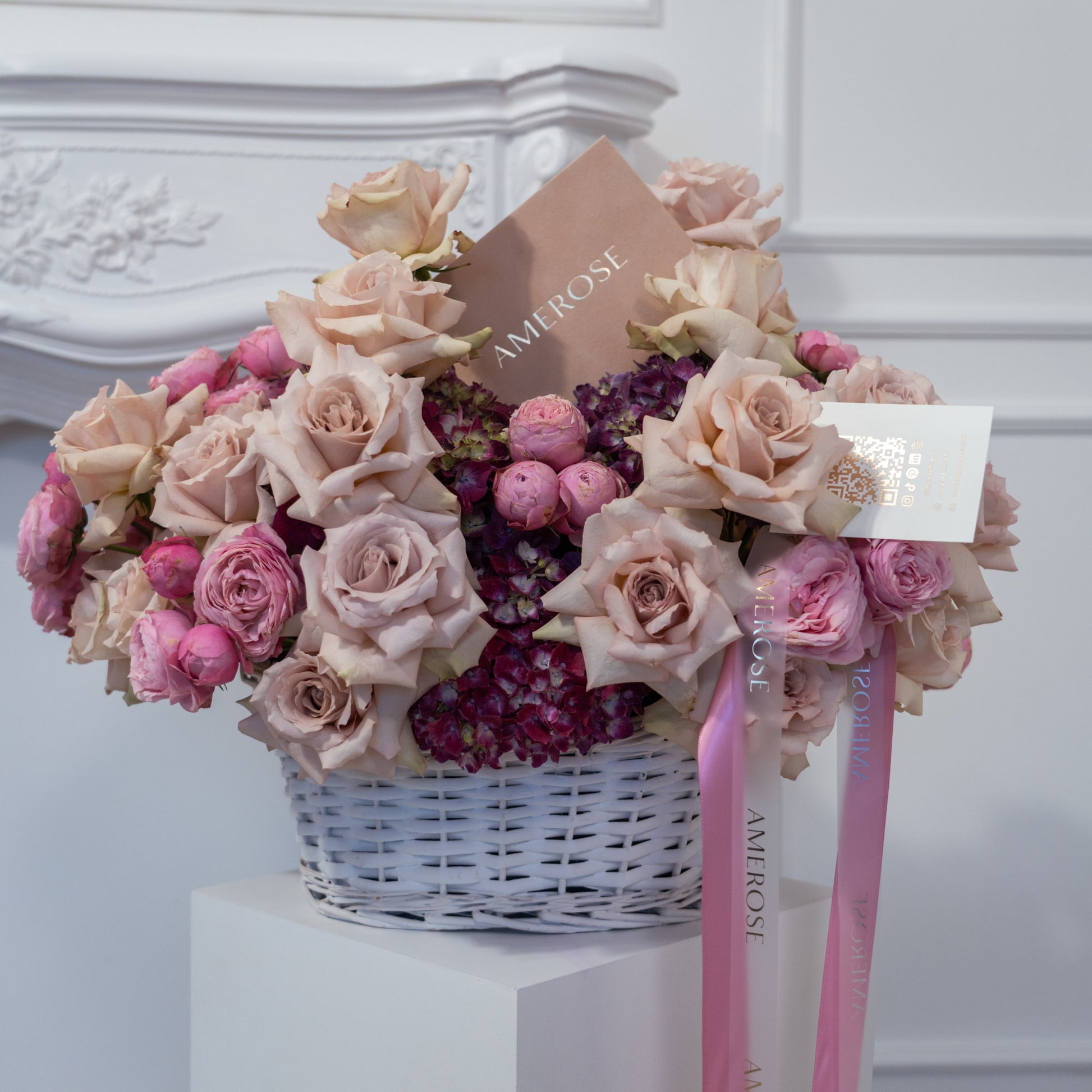 Nude Symphony luxury flower basket by AMEROSE displayed on a white plinth — blush roses, spray roses and hydrangeas in a white wicker basket with personalised card and satin ribbon, London flower delivery