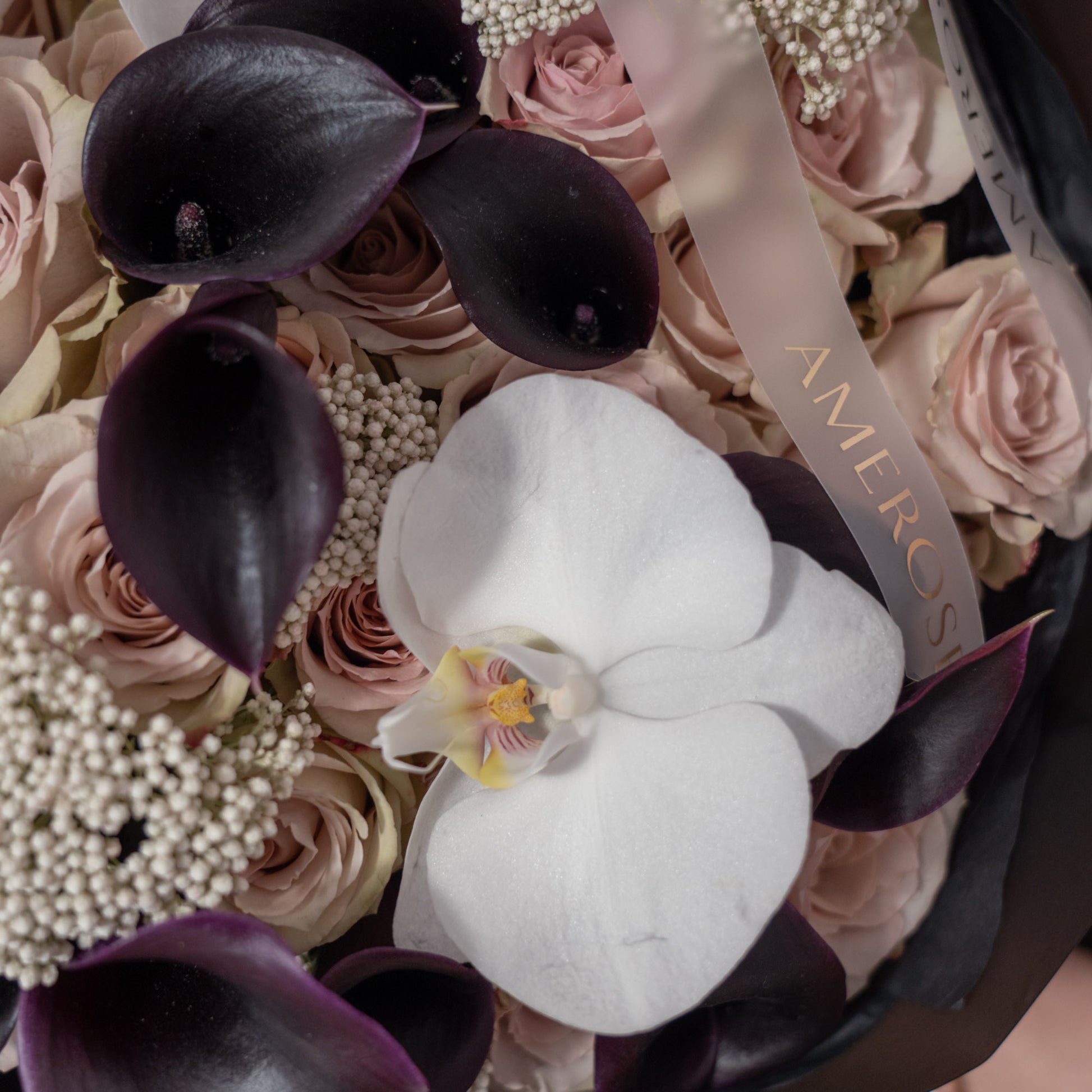 Close-up of deep purple black calla lilies, nude roses, white phalaenopsis orchid and blush rice flowers with AMEROSE ribbon — luxury dark bouquet detail London