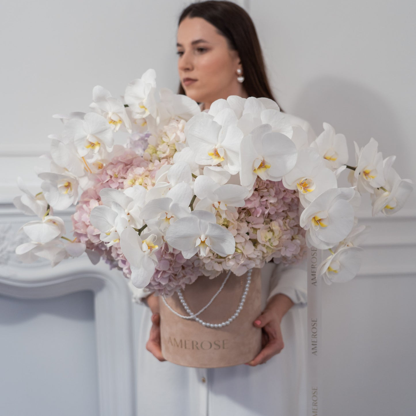 Woman holding a luxury AMEROSE velvet hatbox overflowing with cascading white phalaenopsis orchids and blush hydrangeas with pearl chain handle — statement flower arrangement London