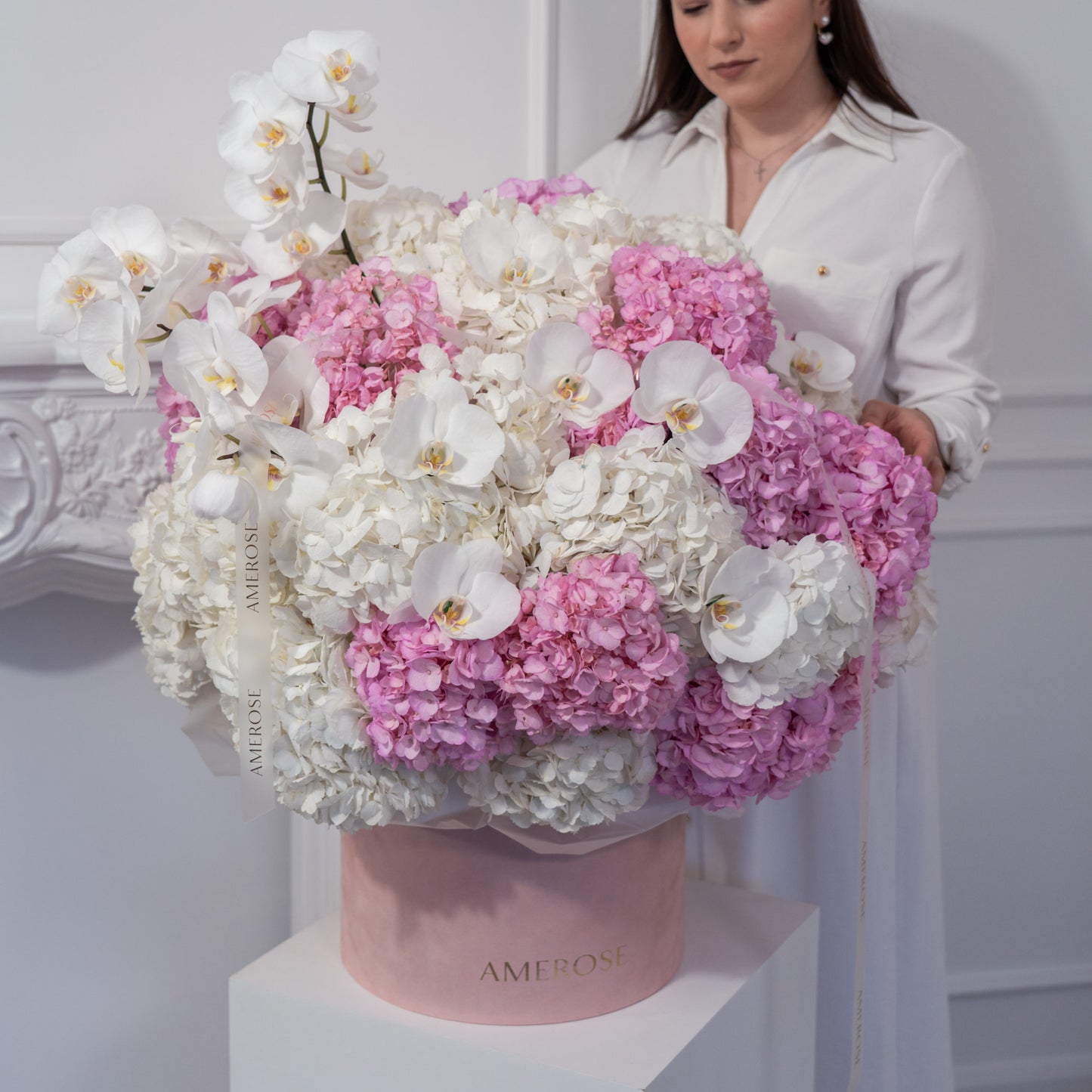 Woman holding an oversized AMEROSE blush velvet hatbox with a cloud of white and pink hydrangeas and orchids on white plinth — statement luxury flower delivery London