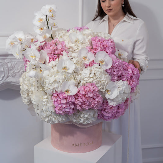 Woman holding an oversized AMEROSE blush velvet hatbox with a cloud of white and pink hydrangeas and orchids on white plinth — statement luxury flower delivery London