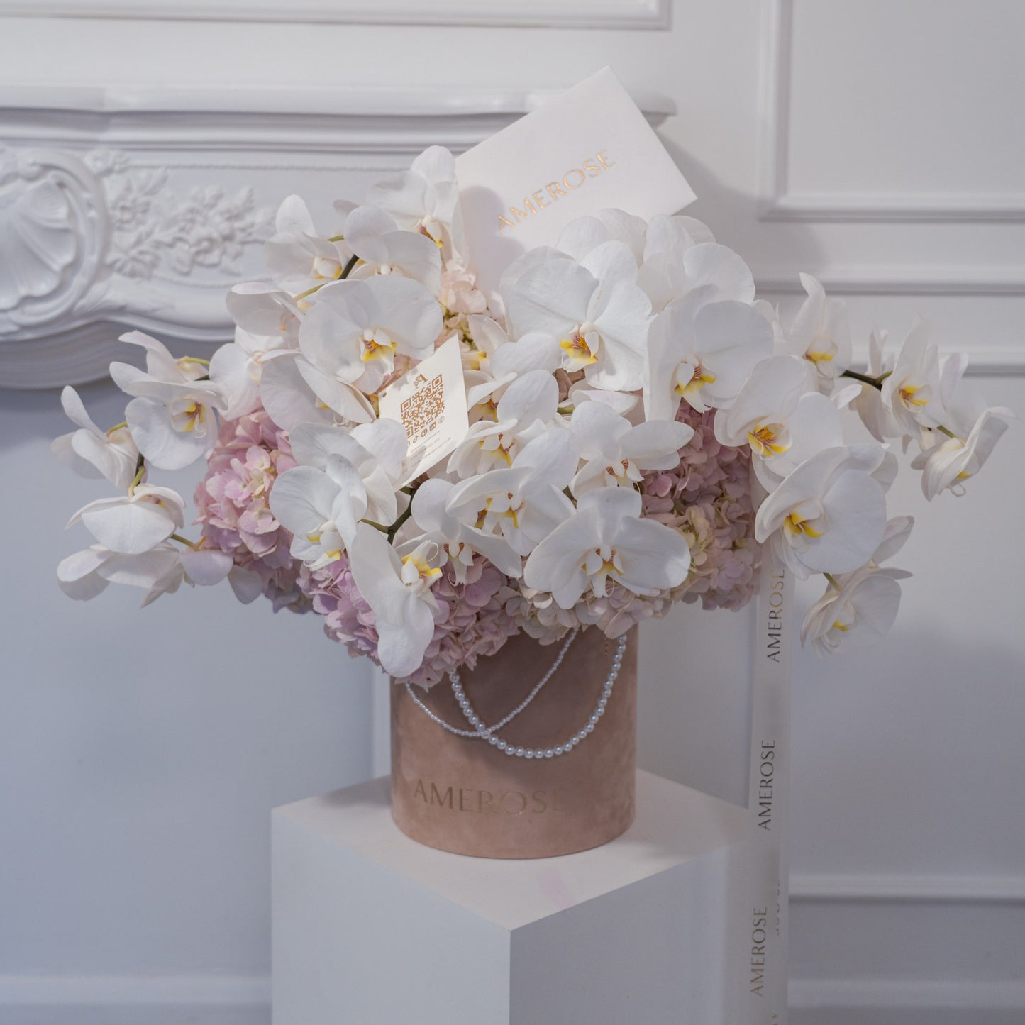 AMEROSE Orchid Couture luxury velvet hatbox with cascading white orchids and blush hydrangeas displayed on white plinth with pearl handle and branded card — luxury flower delivery London