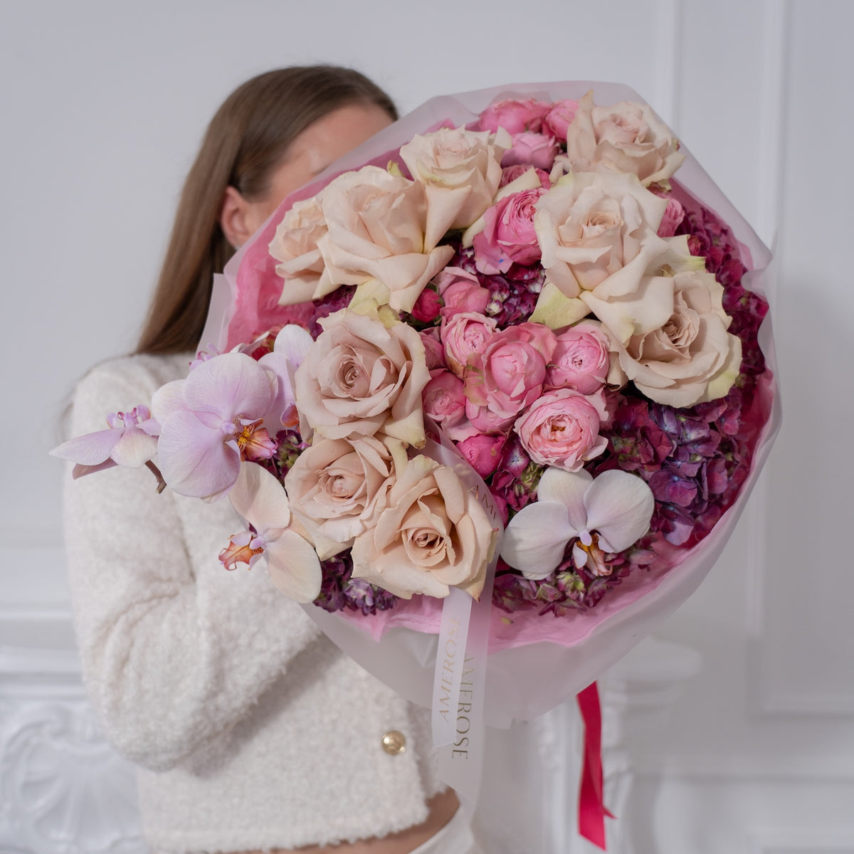 Woman in white bouclé jacket holding a large luxury bouquet of mocha roses, pink spray roses and lavender orchid wrapped in pink film with AMEROSE ribbon — premium flower delivery London