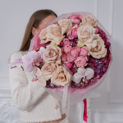 Woman in white bouclé jacket holding a large luxury bouquet of mocha roses, pink spray roses and lavender orchid wrapped in pink film with AMEROSE ribbon — premium flower delivery London